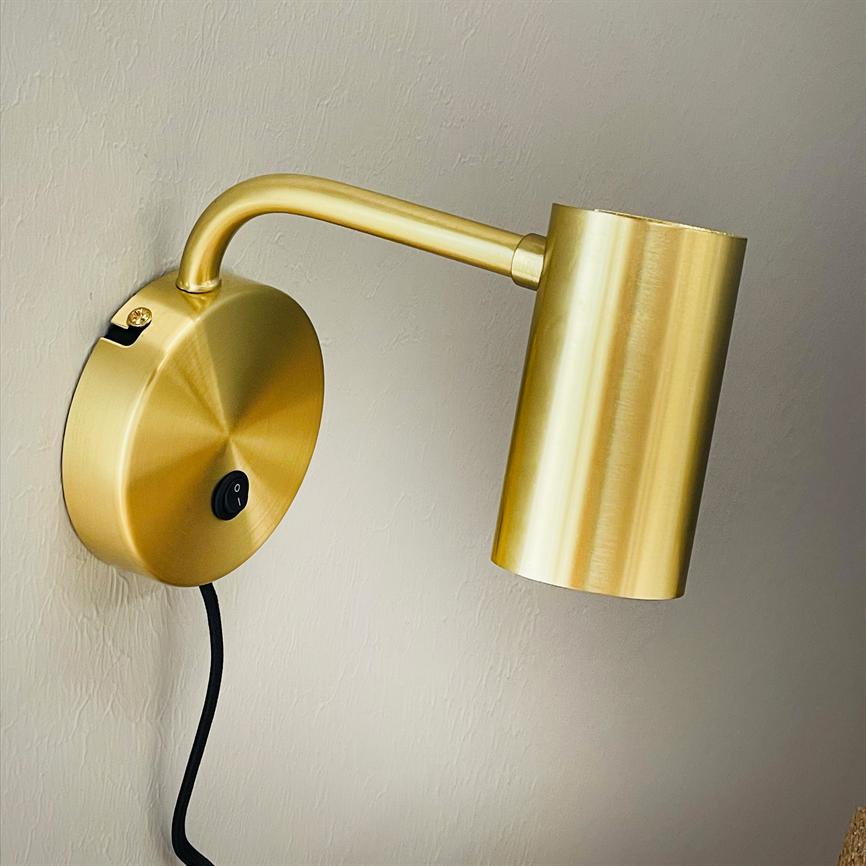 Gold colored wall lamp mounted on a light wall, arm curved out and cylindrical shade directed downwards; black cord hangs freely, on/off button visible on round wall base.