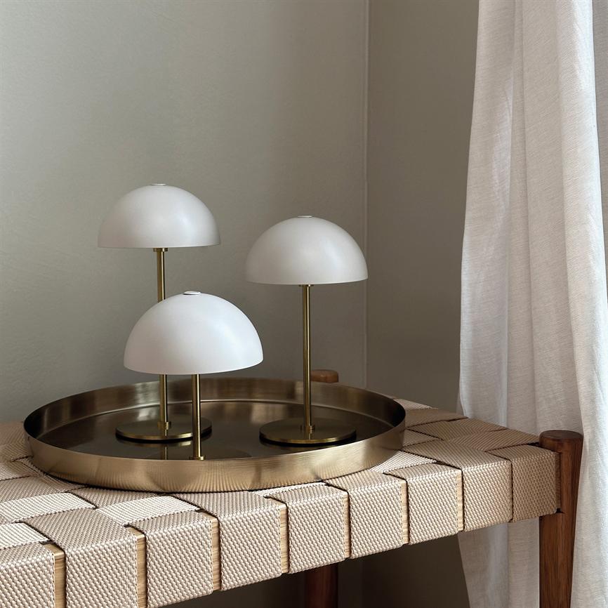 Three white mushroom-shaped table lamps stand on a round brass dish on a light beige wicker bench in a quiet corner by a bright curtain.