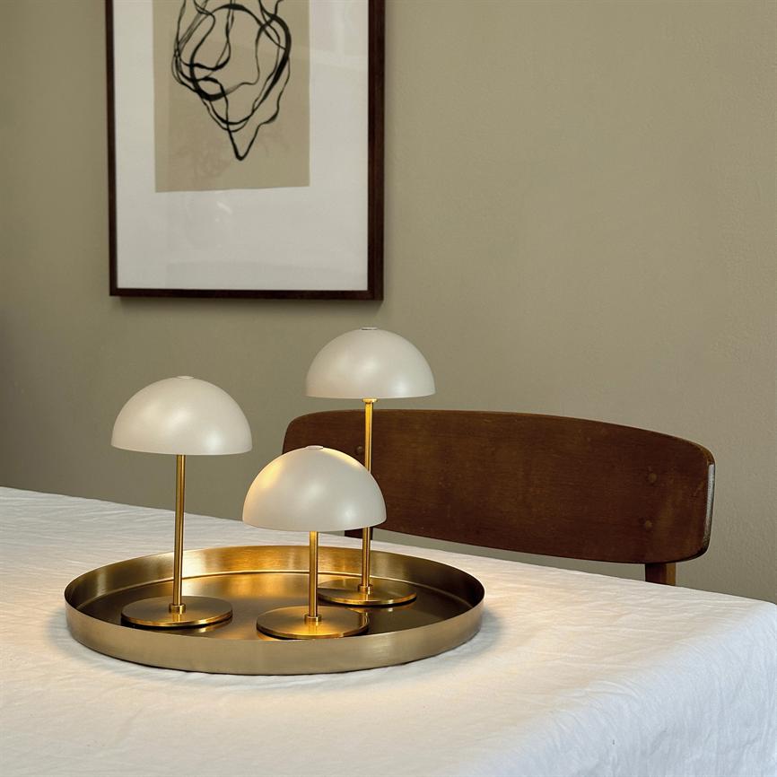 Three small, white dome table lamps stand on a round brass tray; a lamp is lit, casting warm light on a white, dew-covered table in front of a wooden chair and a framed abstract poster.