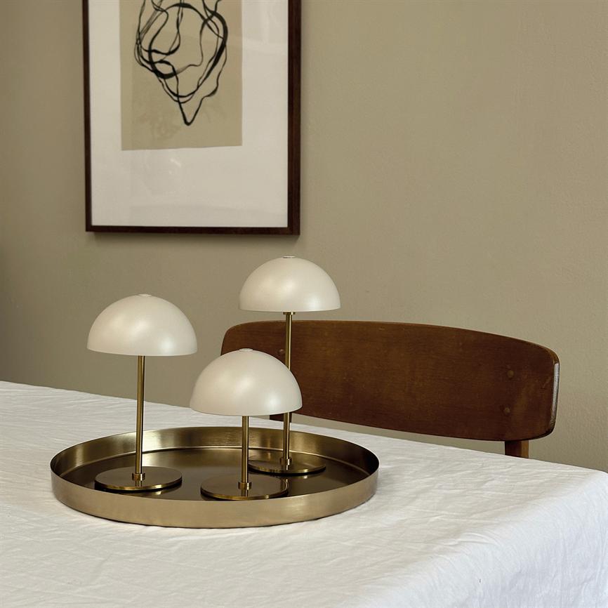 Three small white dome lamps stand on a round brass tray placed on a white tablecloth-covered table in front of a wooden chair and a framed abstract drawing on a beige wall.