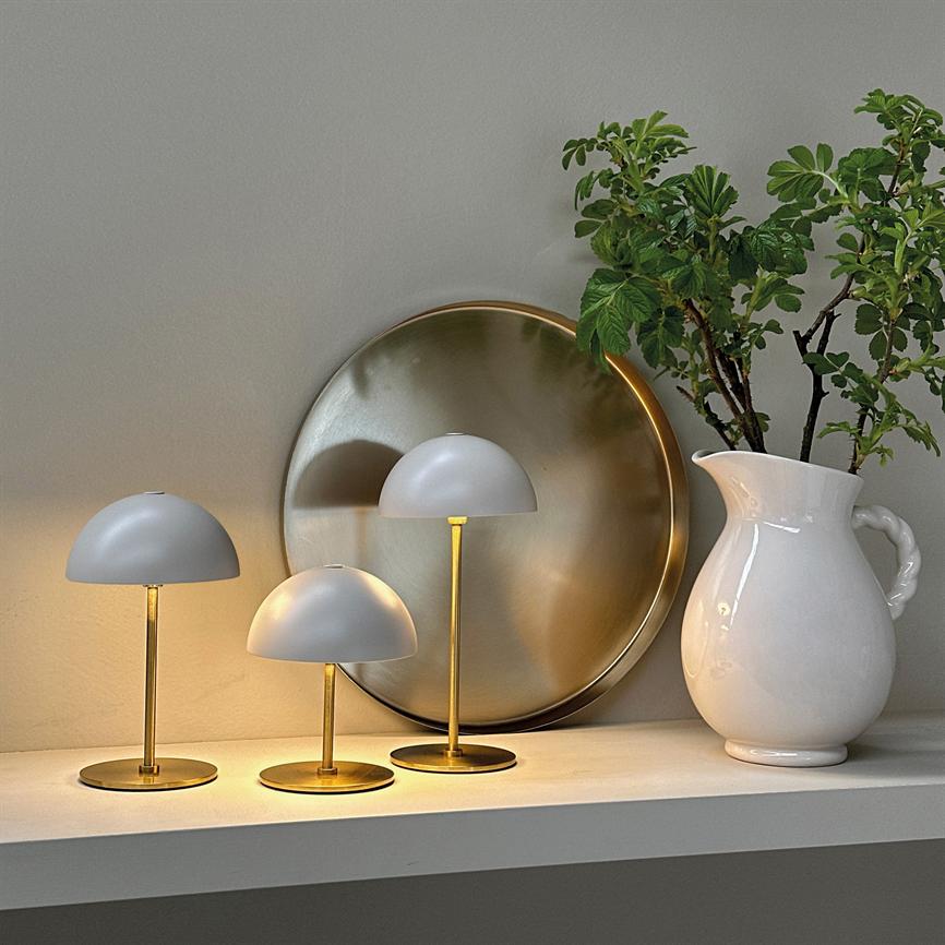 Three small white lampshades are lit on a white shelf; golden metal stands cast warm light against a large brass tray, next to a white pitcher with green branches.