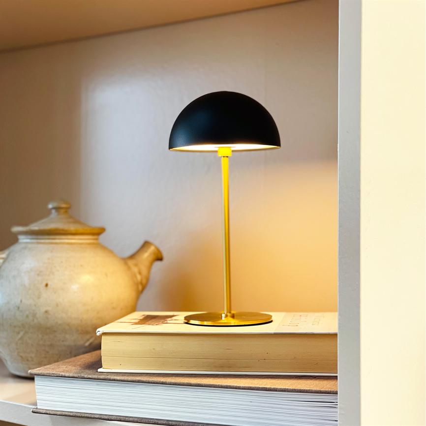 Table lamp with black hemispherical lampshade illuminates warm yellow tones; it sits on a stack of books on a shelf by a rustic ceramic teapot in a soft, warm corner.