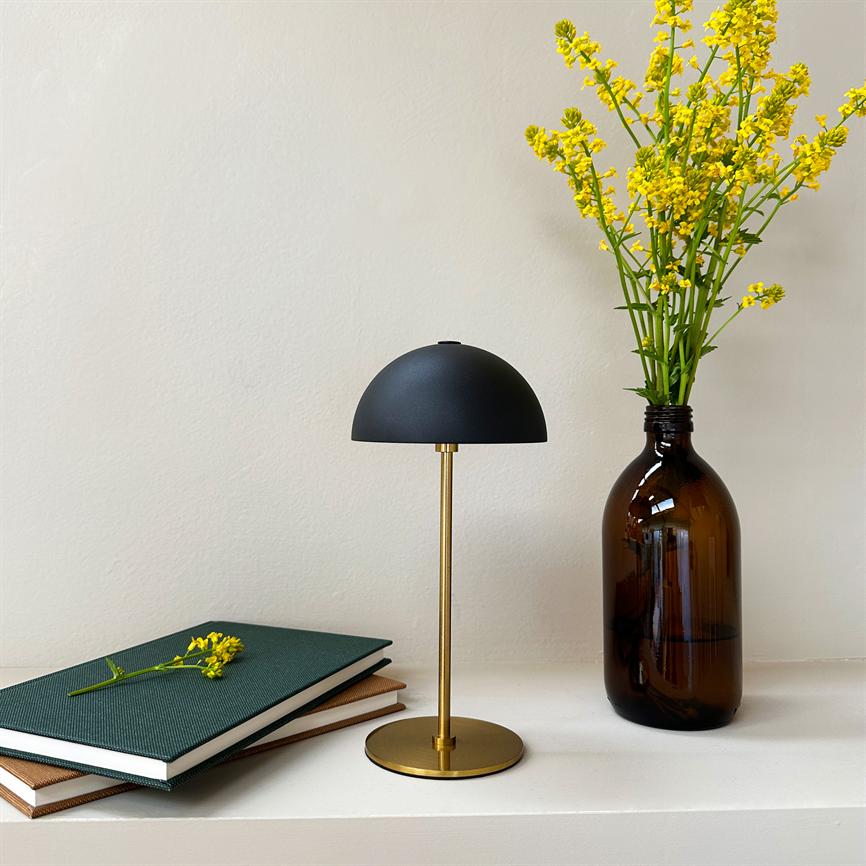 A small table lamp with a black dome and golden base stands on a white shelf between two books and a brown glass bottle with yellow flowers in front of a light wall.