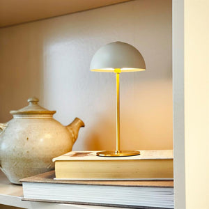 Table lamp with round shade glows softly and warmly, standing on a stack of books in a shelf, with a beige ceramic teapot placed next to it.