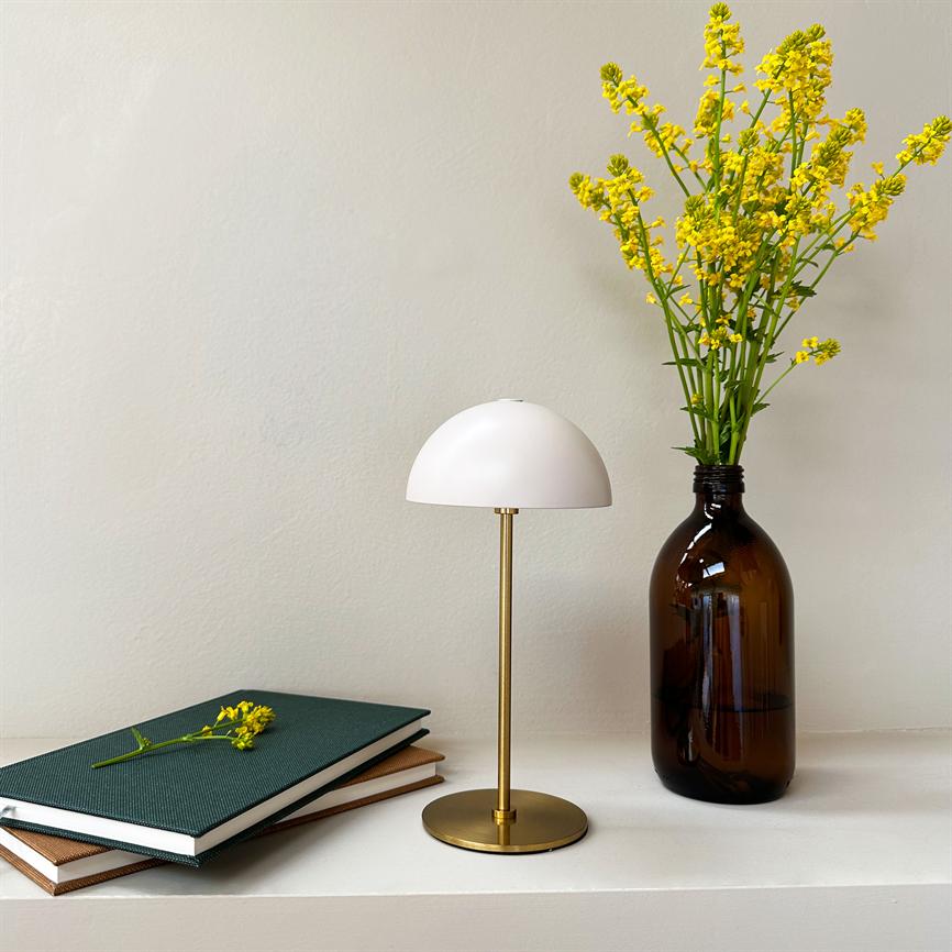 Table lamp with white dome and brass base stands between a stack of notebooks and a brown glass bottle with yellow flowers on a white shelf in front of a light wall.
