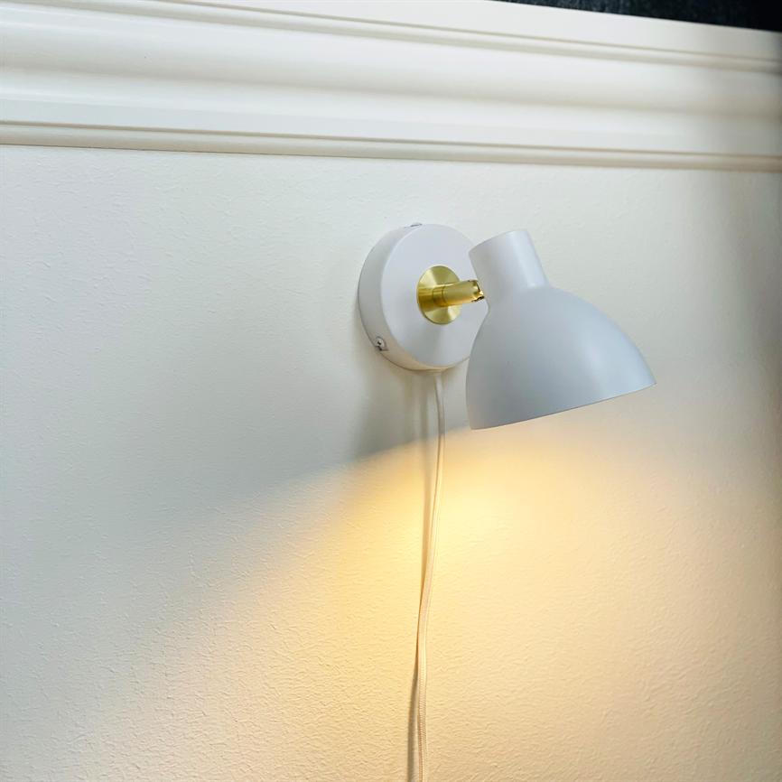 Wall lamp mounted on wall, warm downwards; white shade with gold-coloured arm, round wall base and visible fabric cord hanging along a light wall under profile molding.