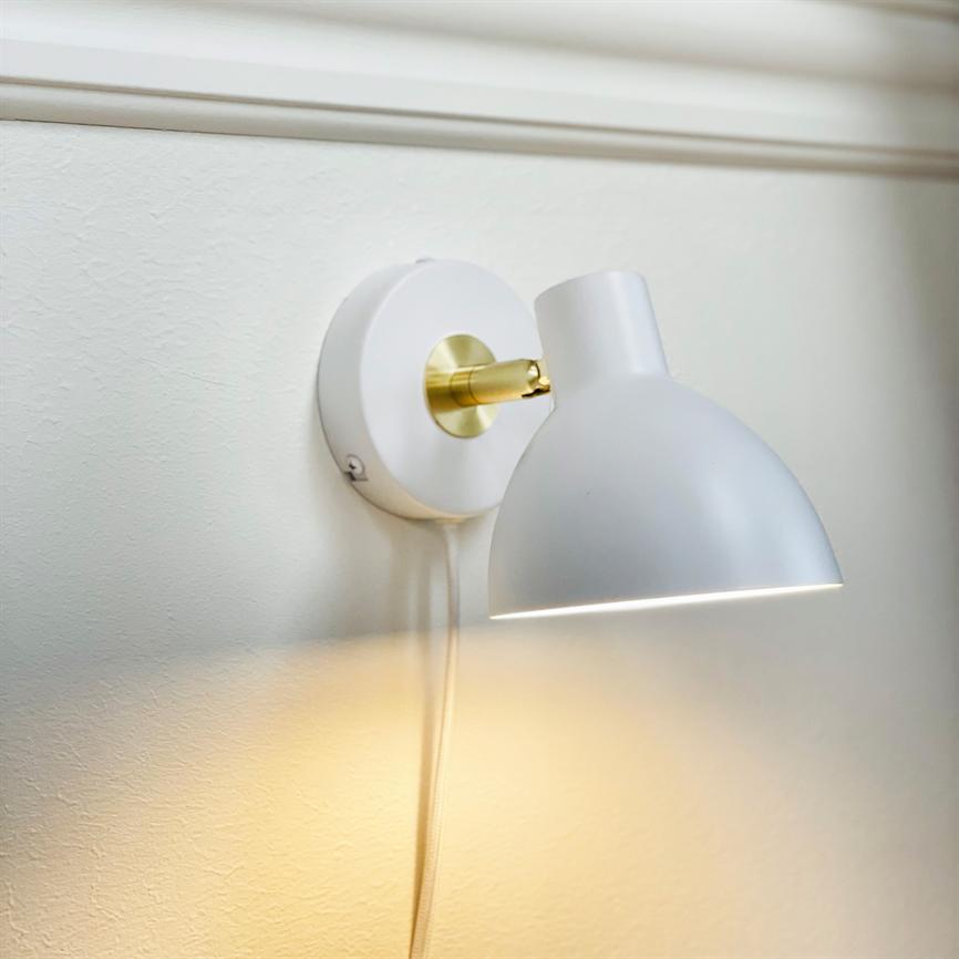A white wall lamp mounted on the wall, turned slightly downwards, emits soft warm light; brass brackets connect screen to round wall plate, and cable hangs down in front of a cream-colored wall with molding.