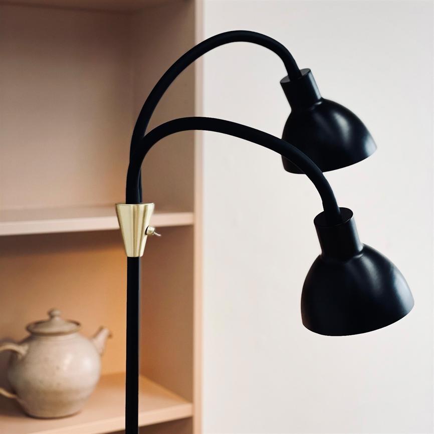 Floor lamp with two curved arms carries two black metal shades, placed in front of a light wall and next to a beige shelf with a ceramic teapot.