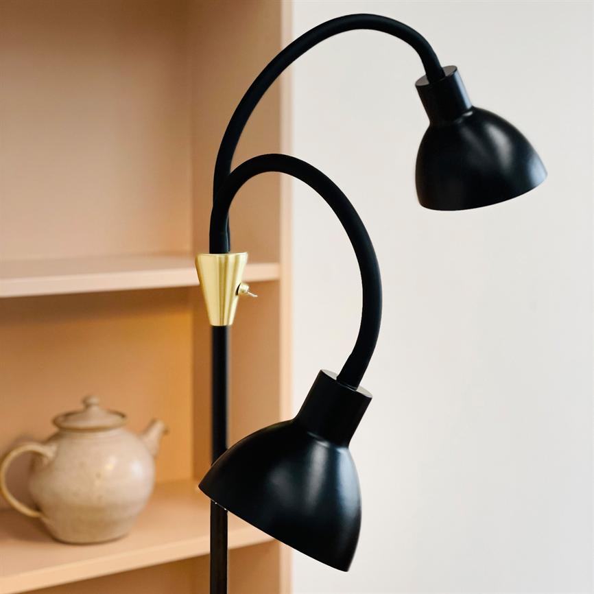 Black floor lamp with two curved arms and matte shades, with a brass detail and small rocker switch; it is switched off by a light wooden bookcase with a beige teapot on the shelf.