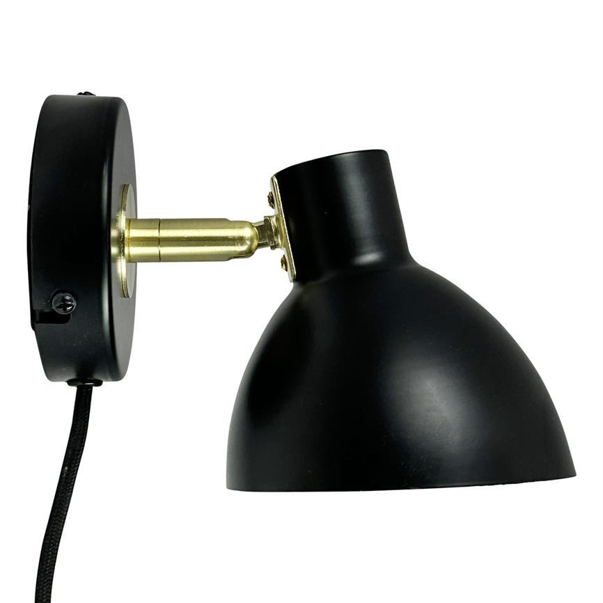 Black wall lamp tilts its hemispherical shade downwards on a brass arm, mounted on a round wall bracket against white background with black fabric cord.