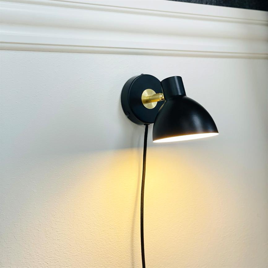 Black wall lamp casts warm, defined light downwards; mounted on a round brass bracket; a black fabric cord hangs vertically against a white wall with decorative molding at the top.
