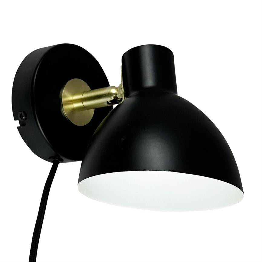 Black wall lamp with round shade, mounted on a round wall bracket; the lampshade is angled downwards via a brass joint, in front of a uniform white background. Black fabric cord hangs down.