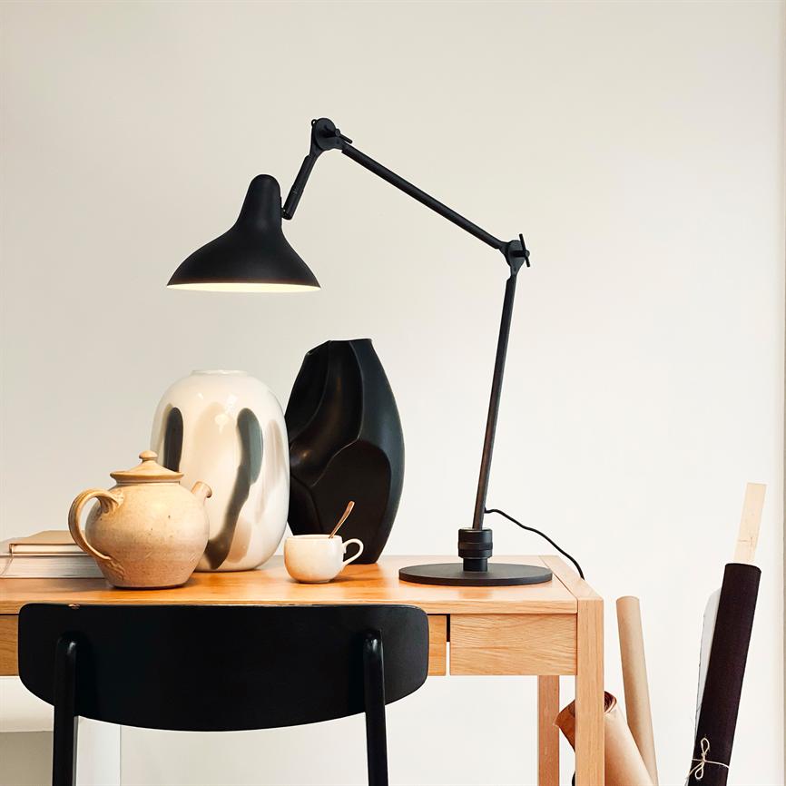 A black adjustable desk lamp shines above a wooden table; it illuminates ceramic vases, a teapot and a cup with a spoon, while a black chair and rolling papers stand in a bright, minimalist room.