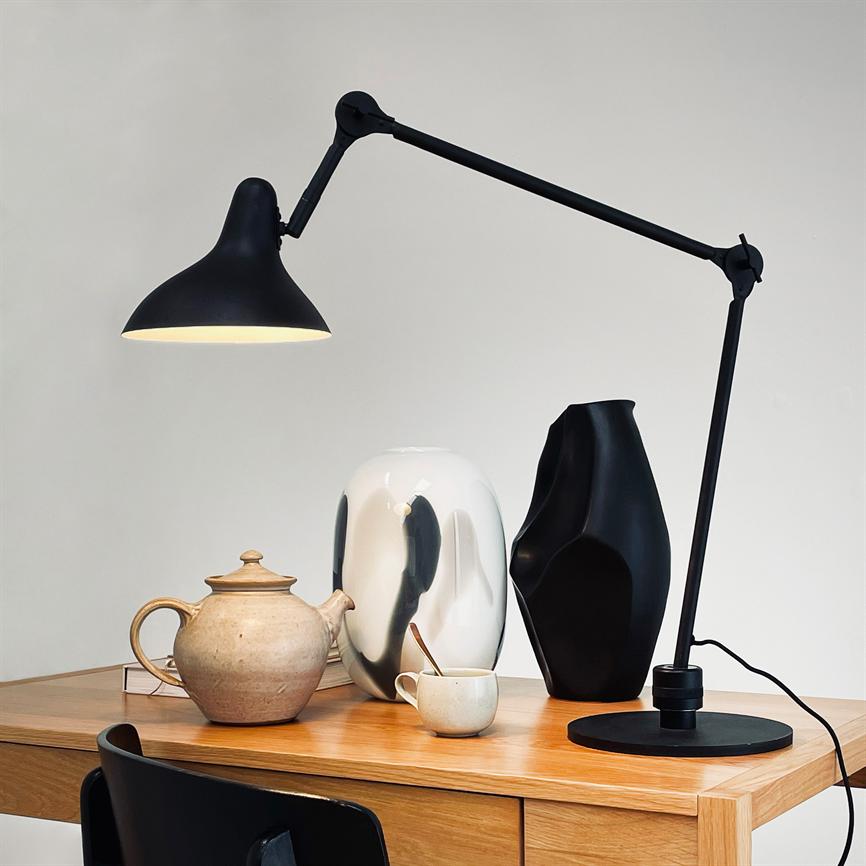 Black, adjustable desk lamp casts warm light over a clay pot and cup on a light wooden desk, flanked by a white and a black ceramic vase in front of a neutral wall.