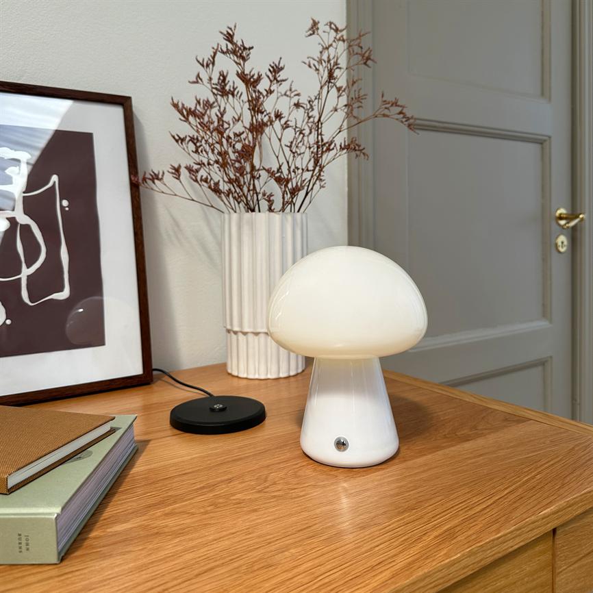 A white mushroom-shaped table lamp glows softly and stands on a wooden cabinet next to a white ceramic vase with dried branches, a framed abstract picture, some books and a closed door.