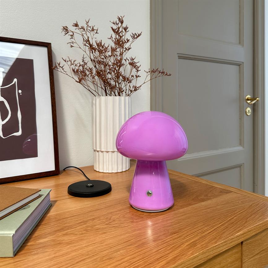 Pink mushroom-shaped table lamp stands on a wooden cabinet, surrounded by a white fluted vase with dried branches, a framed poster and a closed door in the background.