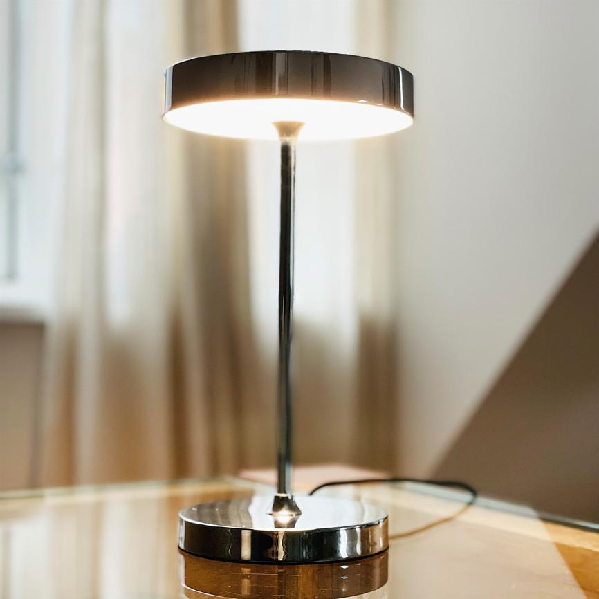 Metal table lamp shines warm downwards; thin rod connects round screen and round, reflective base on a glass table with blurred curtains and a window in the background.