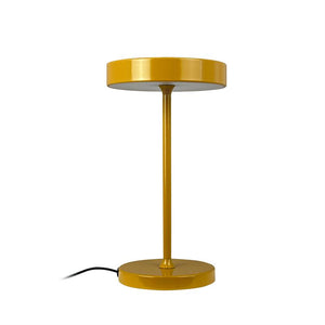 Yellow metal table lamp stands on white background; a slender bar connects the round base and low-profile screen, and a black cord extends faintly to the side.