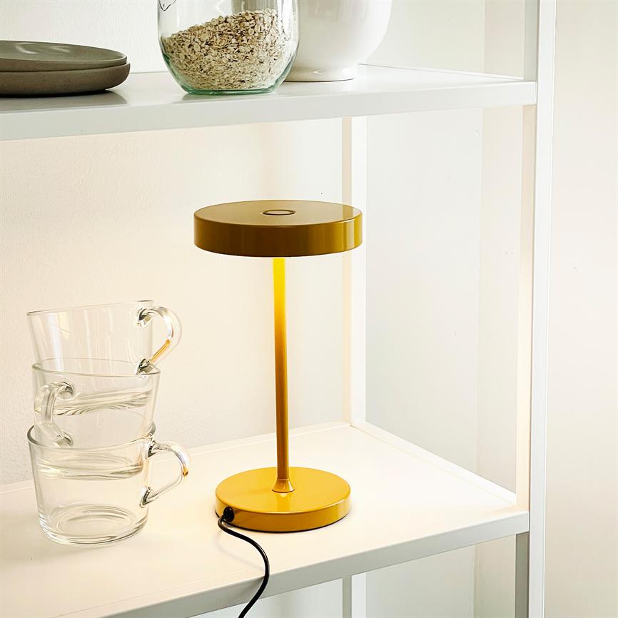 Yellow table lamp is lit on a white shelf; its black cord winds toward the edge; three transparent cups are stacked next to it, and the top shelf holds glass and china containers.