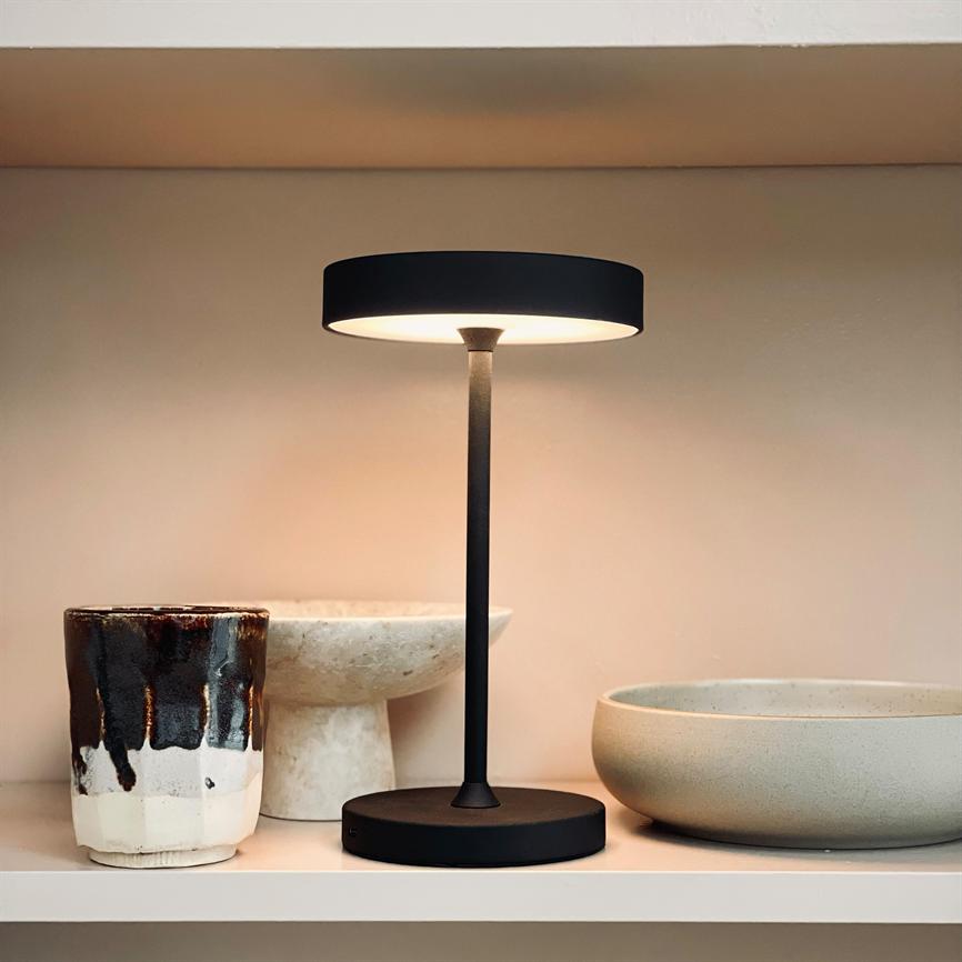 Slender black table lamp stands lit on a shelf, casting soft, warm light; the surroundings consist of ceramic mugs and bowls in neutral tones on a white background.