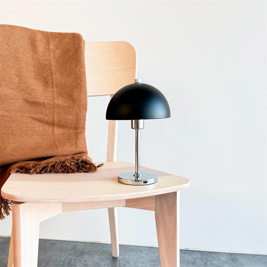 Black table lamp stands on a light wooden chair; a brown carpet lies over the backrest. White wall and gray floor in the background.