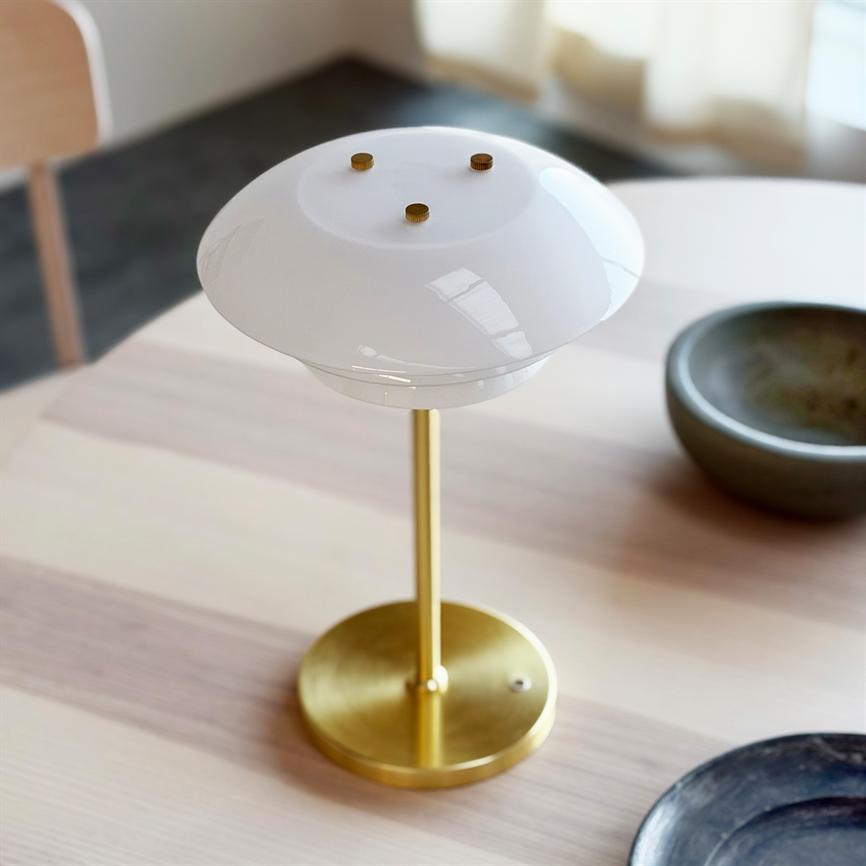 A brass table lamp with a round white glass shade stands on a light wooden table; near a gray ceramic bowl and a black plate in a bright, diffusely lit room.