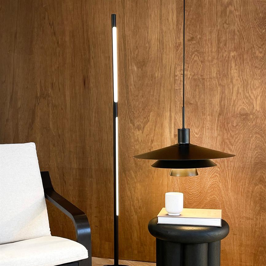 A slim standing LED floor lamp illuminates a corner with a black pendant lamp above a round black side table, where a book and a white mug stand in front of a wood-clad wall.