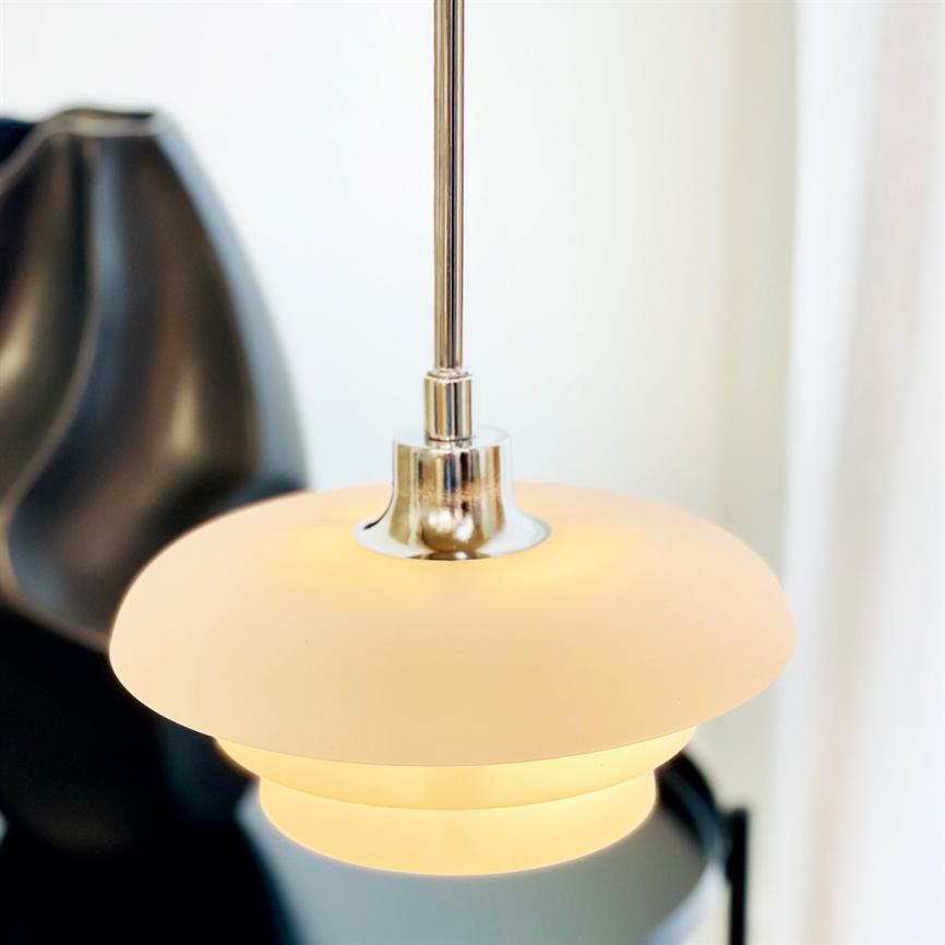 A round white pendant lamp hangs and emits warm light in a bright interior, with a blurred black decorative object in the background and soft white curtains to the right.