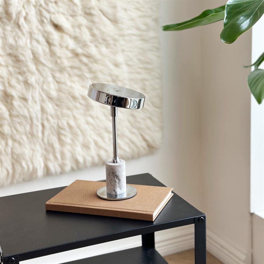 Chrome and marble table lamp stands on a brown notebook, resting on a black metal side table in a bright room with a woolly tapestry on the wall and green plant leaves in the corner.