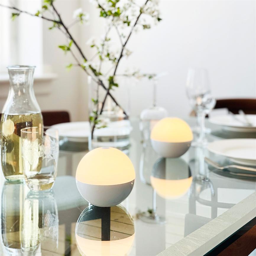 Two round table lamps shine softly and warmly on a glass table surrounded by decanters, glasses, white plates and a vase with flowering sprigs in a bright dining room.