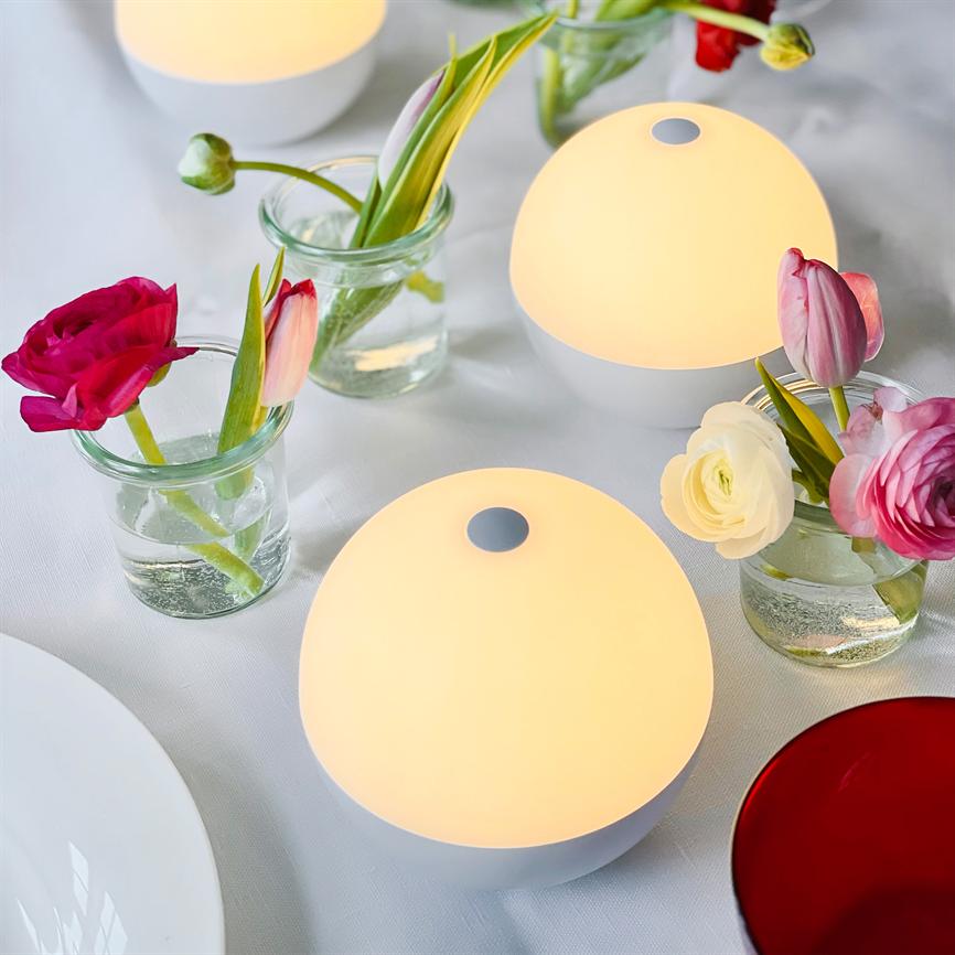 Round white table lamps emit warm, diffused light; they stand among small glass vases with pink and white tulips and buttercups on a white tablecloth as table decorations.