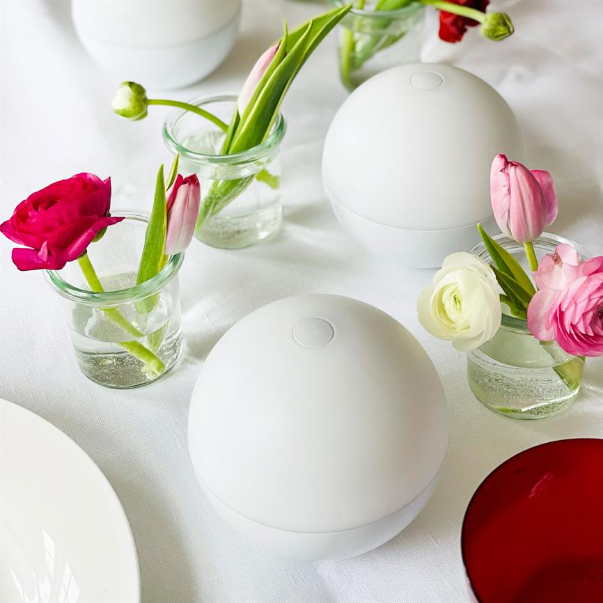Two white spherical units are placed on a white tablecloth, surrounded by small glass vases with pink and white tulips and buttercups, as well as a white plate and a red glass.