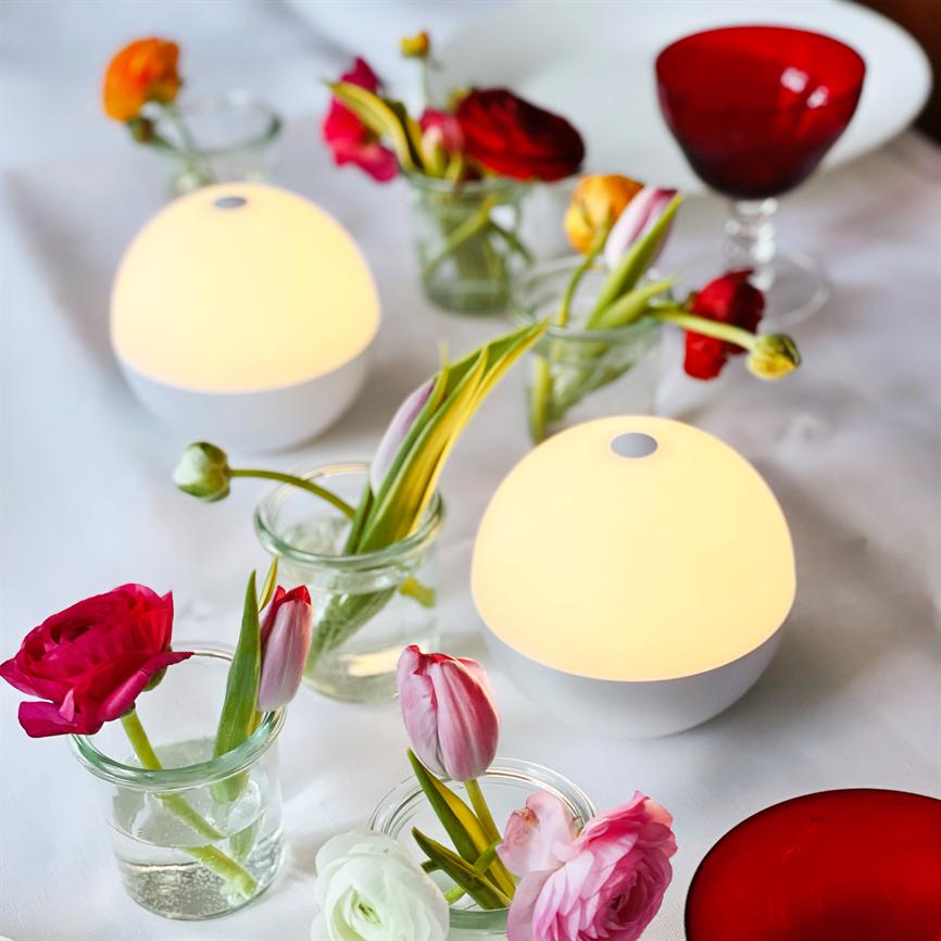 Two round table lamps emit warm, diffused light; they stand among small glass vases with pink, red and white flowers on a white tablecloth next to a red wine glass.