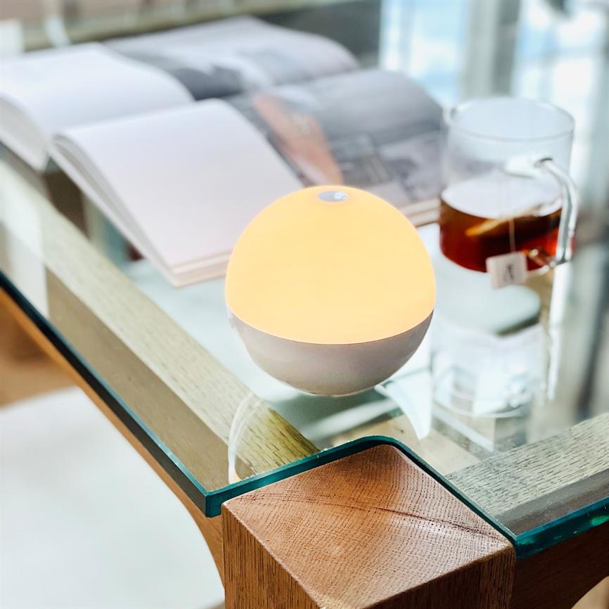 A round, white night lamp emits a warm orange glow, placed on a glass table; next to it is an open book and a glass mug of tea. Text on the tea label: "A".