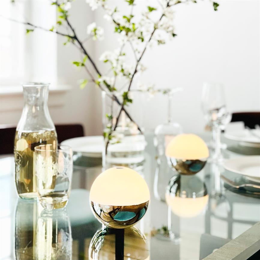Two small spherical table lamps emit warm, soft light; they are reflected in a glass table surrounded by wine glasses, a water carafe, plates and a vase of white flowers in a bright dining room.