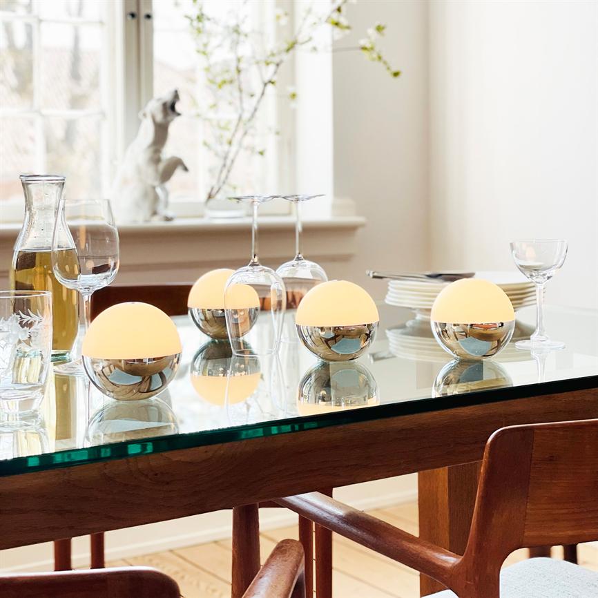Round table lamps stand on a glass table, casting warm light and reflecting between wine glasses, decanters and stacks of plates in a bright dining kitchen by the window.