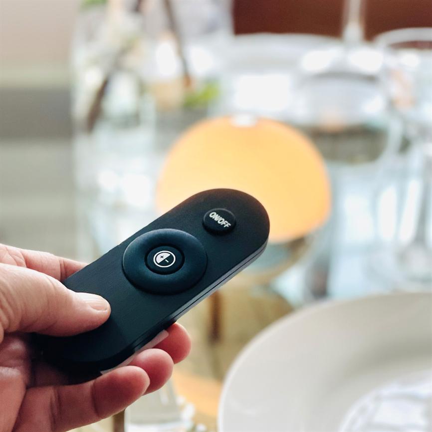 A black remote control is held in the hand and pointed at a warm orange table lamp; glasses and a plate are blurred on a covered table in the background.
 
 Text in the image: "ON/OFF" and "+"