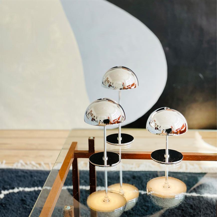 Three chrome lamps mirror the room, placed on rod feet above a glass table; below them are seen warm reflections and a dark carpet, with an abstract black-and-white wall in the background.