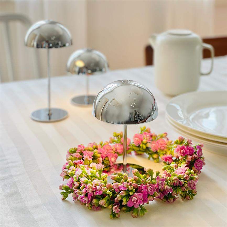 Sølvfarvede, halvkugleformede stande står på en hvid, stribet dug; en krans af pink småblomster omfavner den forreste stand, med stakkede porcelæntallerkener og en hvid kande i baggrunden.