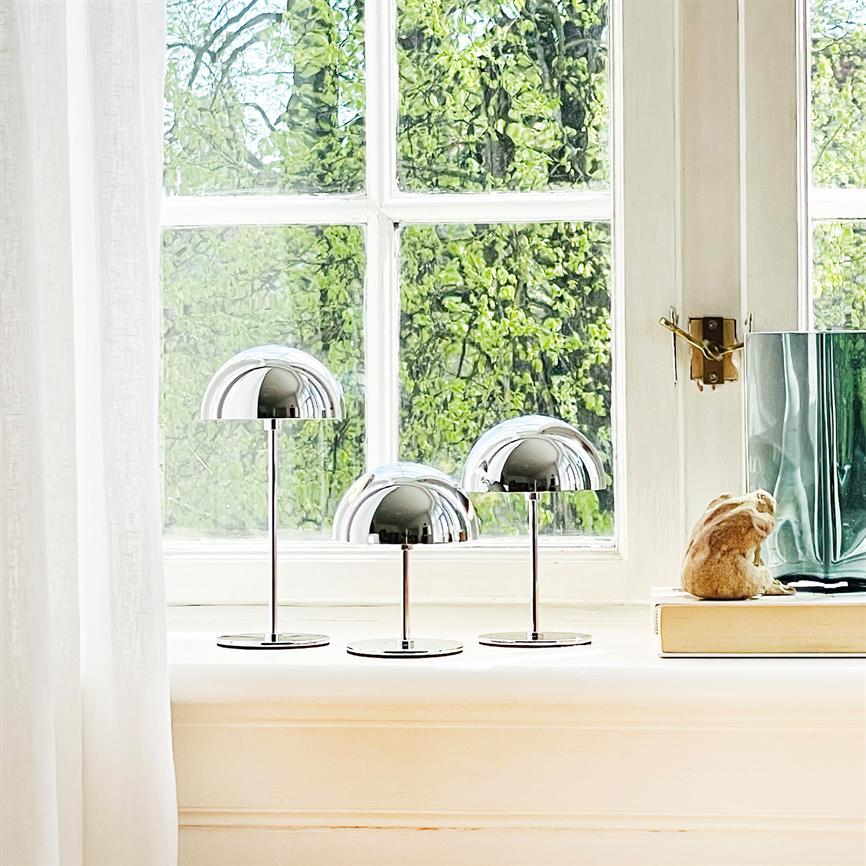 Tre krom-halvkuglelamper står på en hvid vindueskarm og reflekterer lys, foran grønne træer udenfor; et hvidt gardin, en bog, kaninfigur og grøn glasvase ses ved siden af.