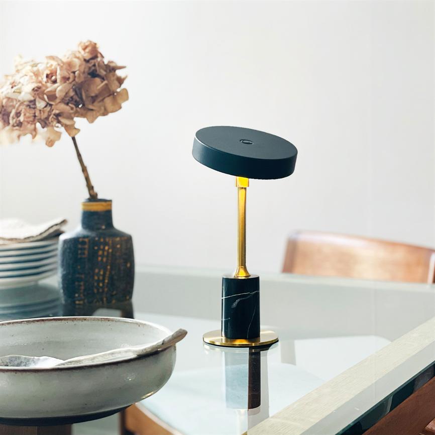 Table lamp stands on a glass table in a bright dining room; gold colored stem and black shade, dried flower in vase, ceramic bowl and stacked plates around.