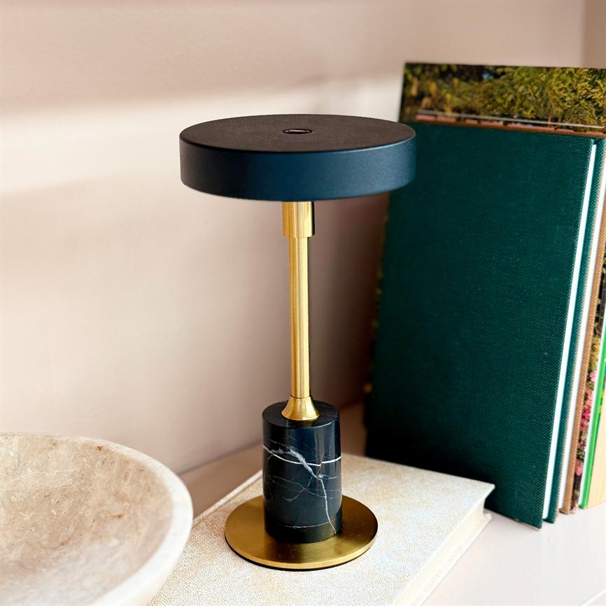 Table lamp stands on a shelf; black round shade and gold colored rod above a black marble base with white veins; flanked by green books on the right and a beige bowl on the left.