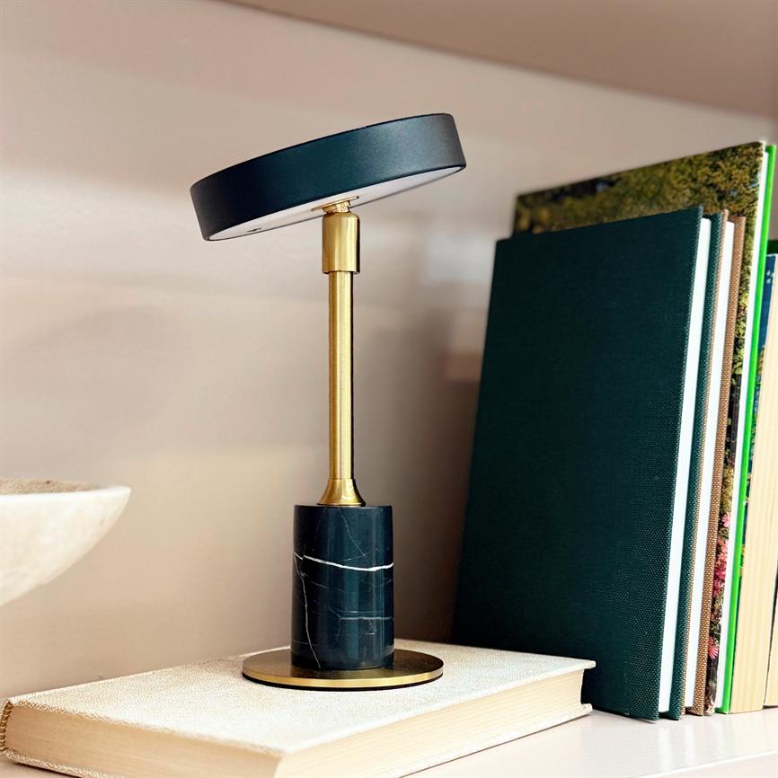 Table lamp with black round shade and brass rod stands slightly tilted on a black marble base, placed on a thick white book on a shelf next to several green bound books.