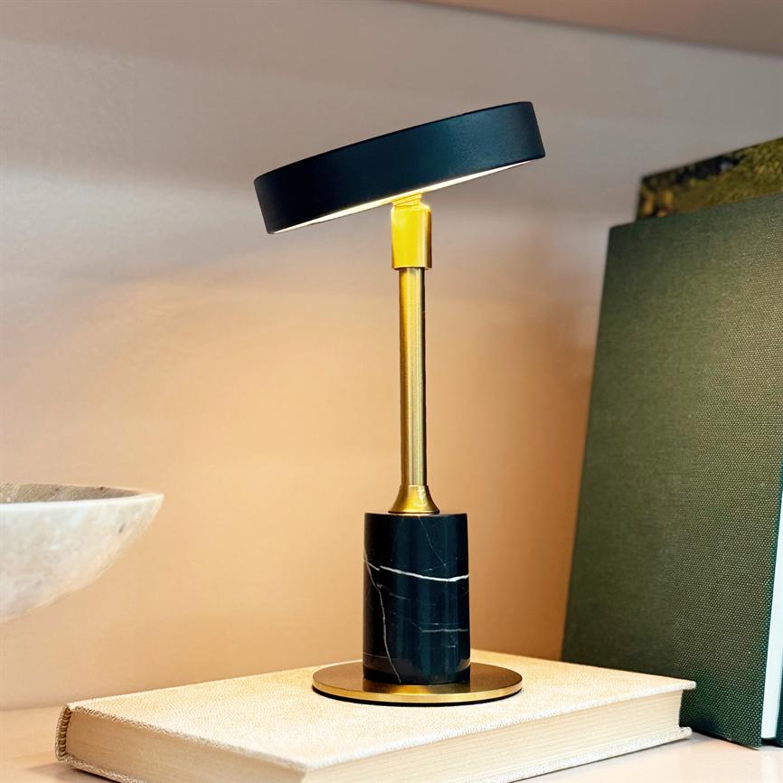 Desk lamp is lit on a closed book, its round black shade tilts slightly and casts warm light against a beige wall; green book to the right and bowl to the left.