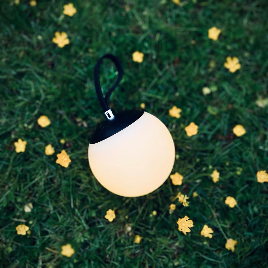 A round, glowing torch with a black top and carrying handle glows softly, resting on green grass surrounded by small yellow flowers.