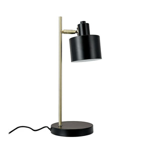 Table lamp with black cylindrical shade, tilted downwards, mounted on a slender brass rod and round black base; visible black wire on uniform white background.