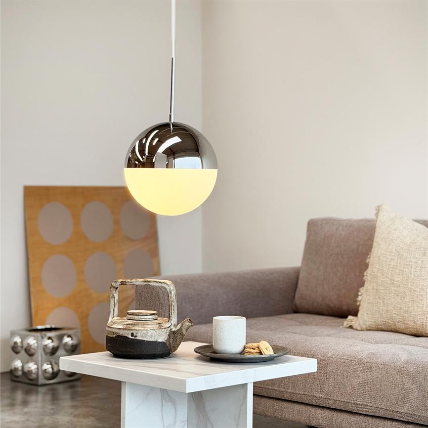 A round pendant lamp hangs lit above a small marble table; on the table is a rustic teapot, cup and cookies. In the background a beige sofa, decorative cushion and decorative panel.
