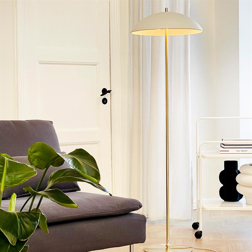 Floor lamp is switched on and casts warm light in a calm living room environment by a gray sofa, green house plant, white door and rolling table with book and vases. Text: "THE TOUCH".