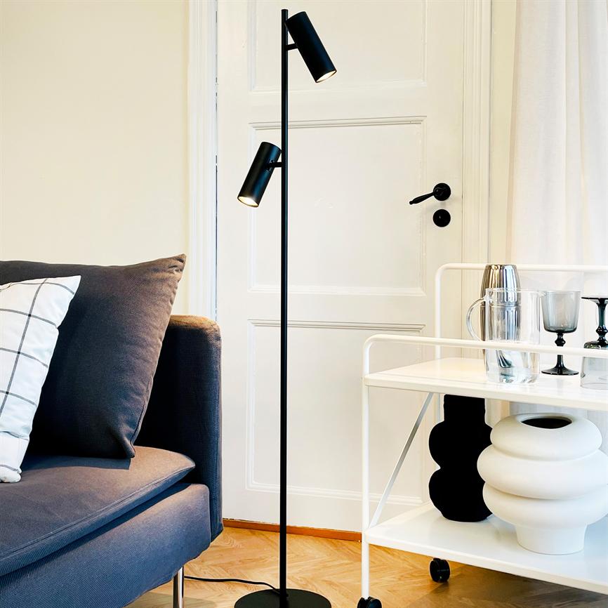 A slim, black floor lamp shines down by a white door, between a blue-grey sofa with a patterned cushion and a white trolley with glasses, jugs and decorative vases.