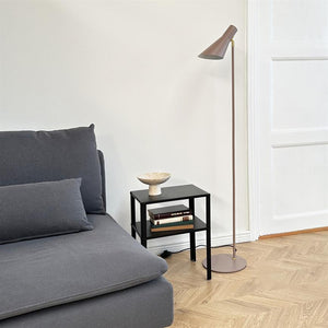 Gray sofa is located on the left; a small black side table bears a bowl and a stack of books; a slim floor lamp stands next to it in a bright room with a parquet floor.